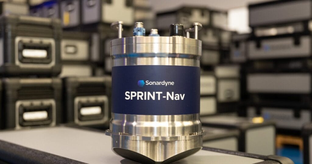 A cylindrical, silver-colored Sonardyne SPRINT-Nav device with a blue label, sitting on a grey surface with a blurred background of industrial cases.